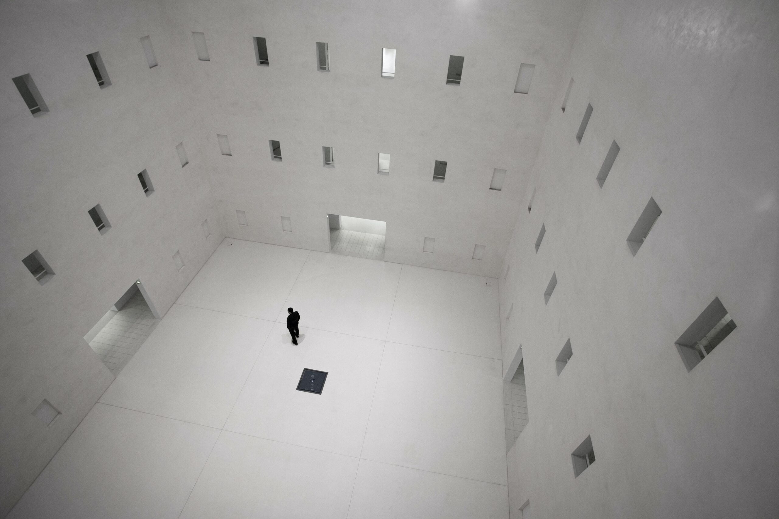 Man standing alone in empty, white room, facing windows with lights on and off